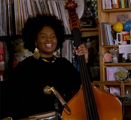 Endea Owens smiles as she plays her bass on the NPR Tiny Desk concert set, with bookshelves in the background.